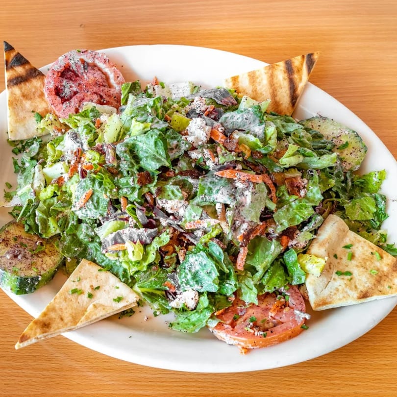 A Fanoos Grill salad with lettuce, tomatoes, cucumbers, feta, and pita bread on a white plate.
