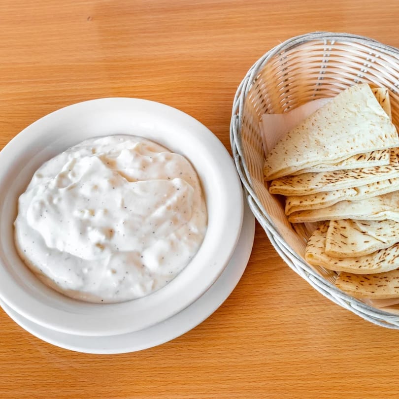 A bowl of creamy white dip with black specks next to a basket of pita bread on a wooden table.